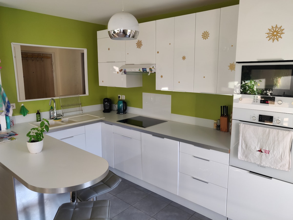 A modern kitchen features sleek white cabinetry and a light green accent wall. A countertop with two bar stools is positioned near a large window, providing natural light. Appliances include an electric stove, oven, and coffee maker, along with a small potted plant for decoration.