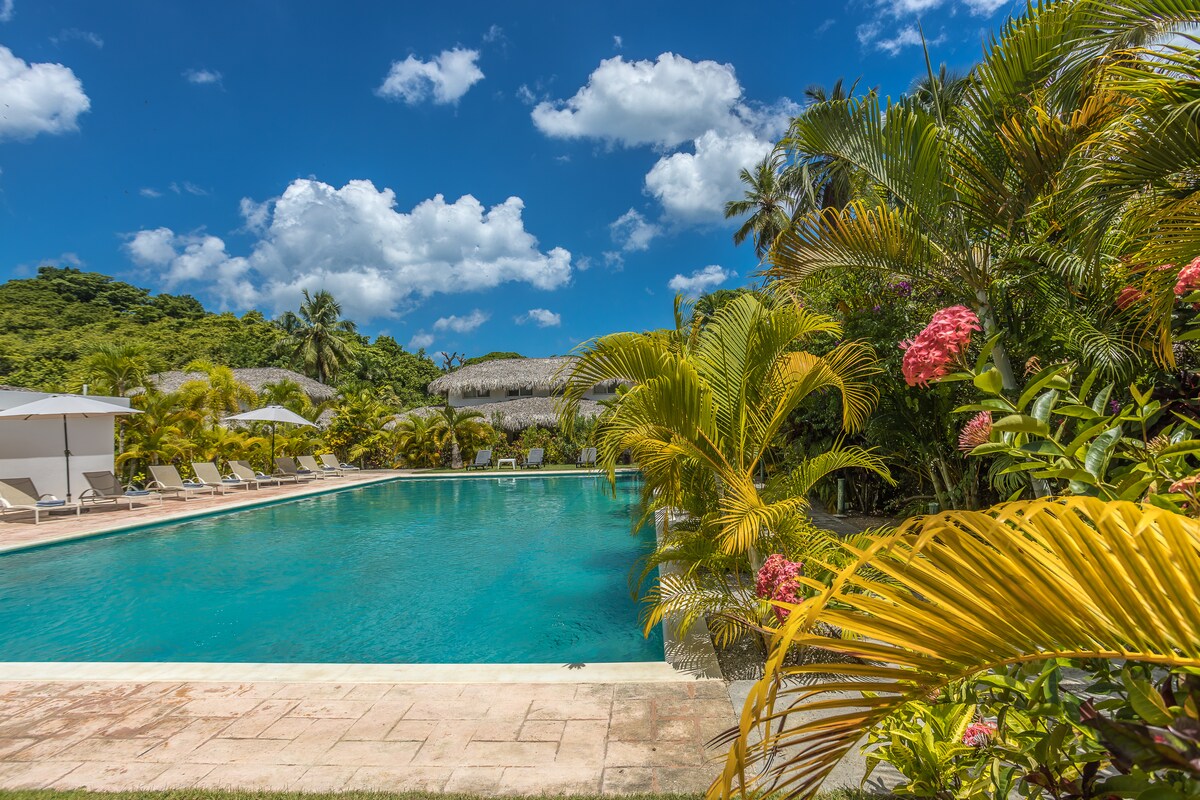 A spacious communal pool is surrounded by lush tropical gardens, featuring various palm trees and vibrant flowers. Lounge chairs are arranged along the water's edge, and the backdrop includes hills and thatched-roof structures under a clear blue sky with scattered clouds.
