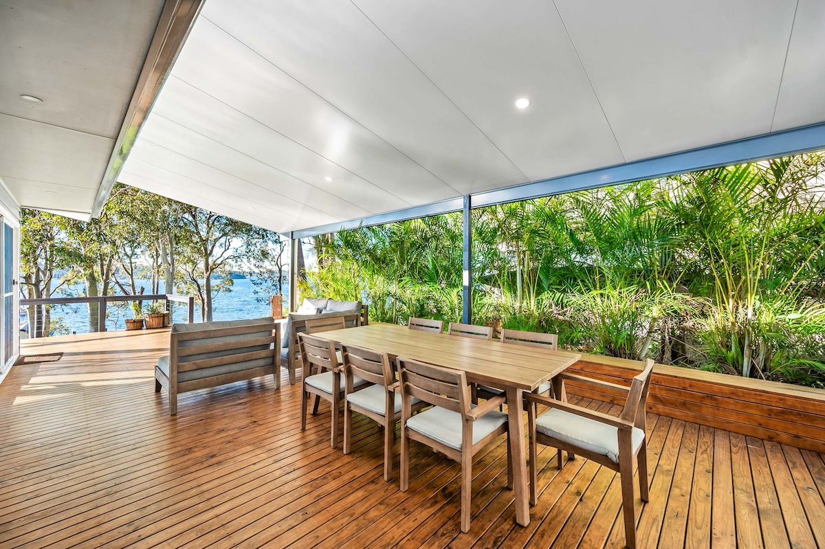 An expansive deck, featuring a large wooden dining table surrounded by eight comfortable chairs, is presented. Lush tropical gardens provide a natural backdrop, while the lake offers scenic views. The roof covers the entire deck for protection from the elements.