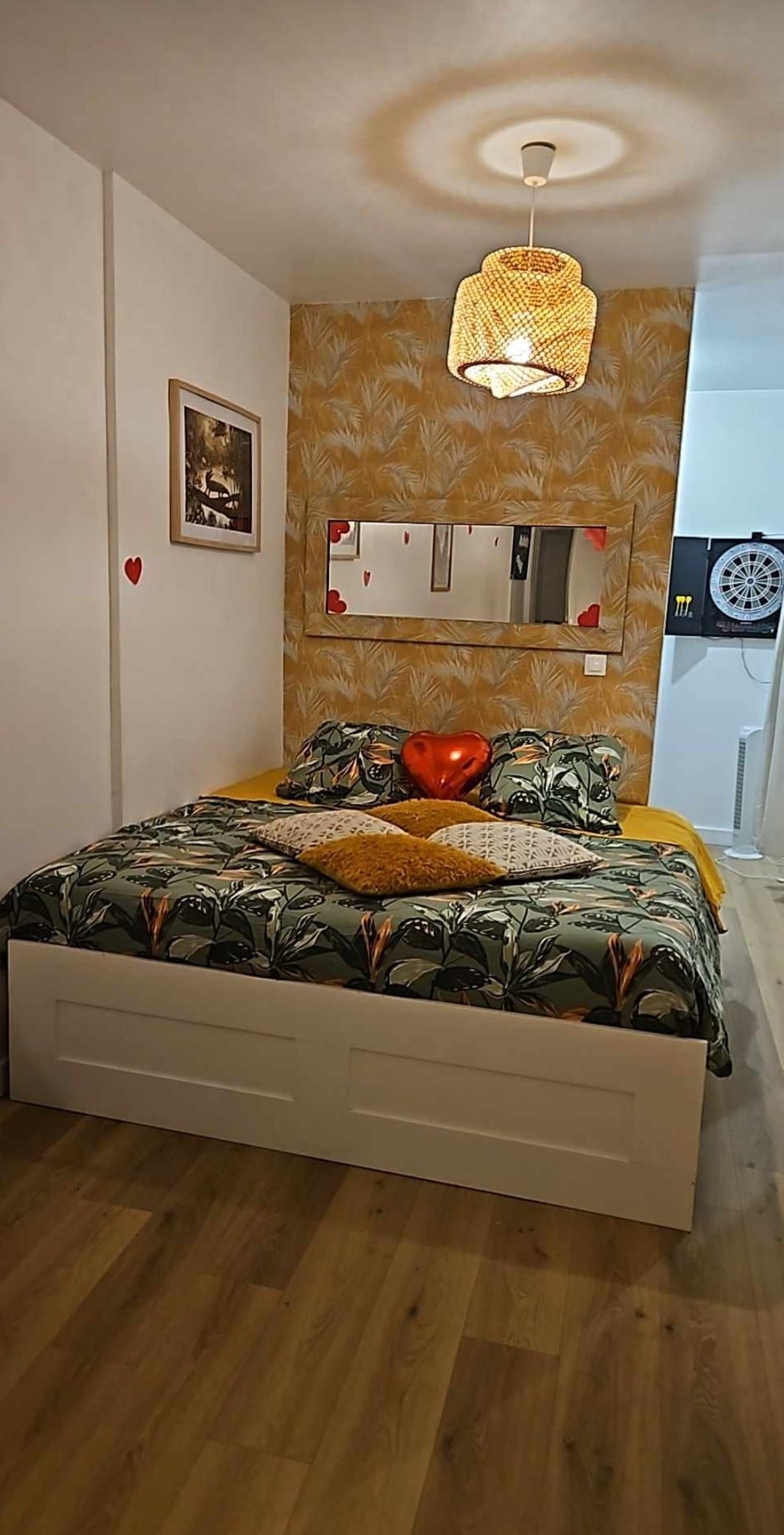 A bright bedroom is presented with a spacious bed featuring a colorful botanical patterned duvet. Decorative pillows in warm tones are arranged on the bed. A rattan light fixture hangs above, complemented by a mirror reflecting the room's cheerful decor.