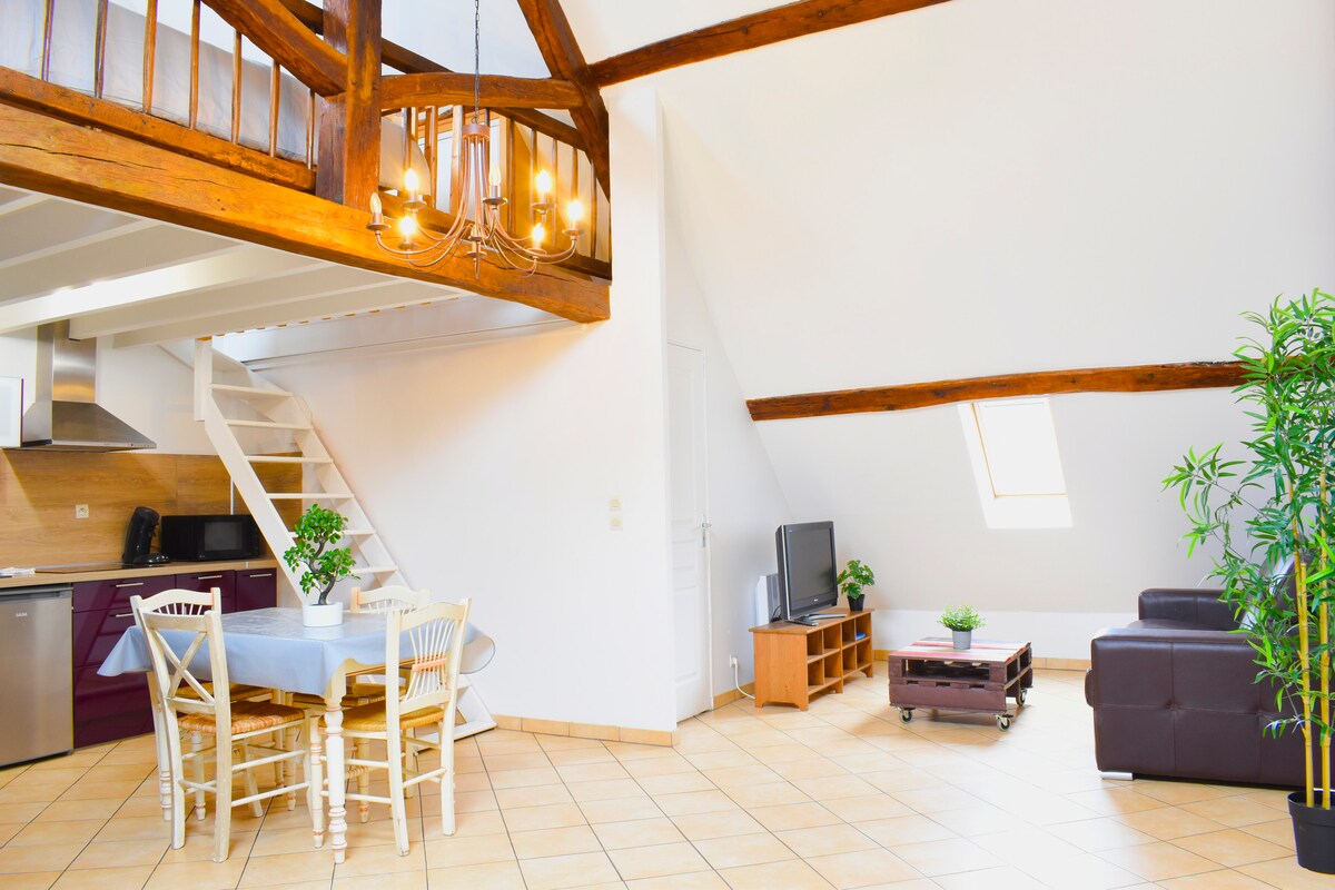 The spacious loft features wooden beams and warm tile flooring. A dining area with a table and six chairs is accompanied by a well-equipped kitchen. A cozy seating area with a dark couch and television is present, alongside a staircase leading to the mezzanine.