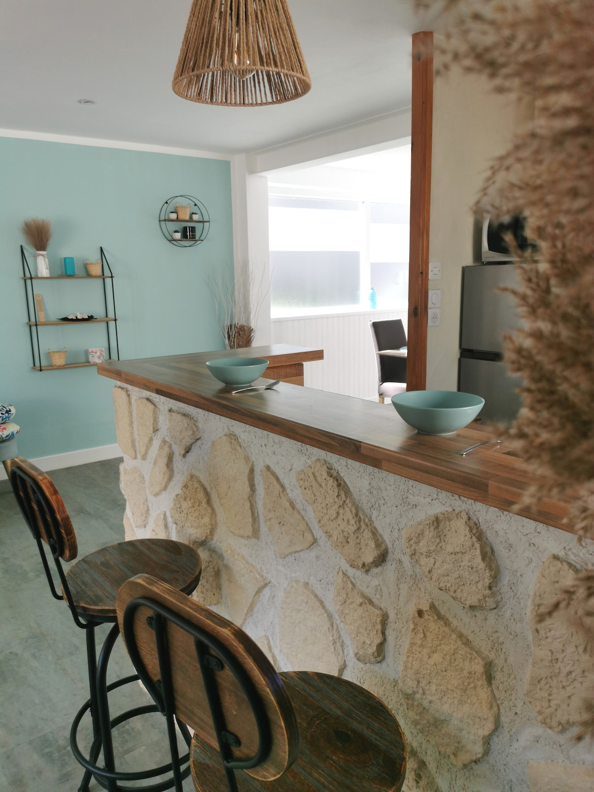 A bar area features three wooden stools positioned at a stone counter, with light blue bowls placed at each seat. A textured wall enhances the space, while decorative items are arranged on a nearby shelf. Natural light filters in, creating a bright and welcoming setting.