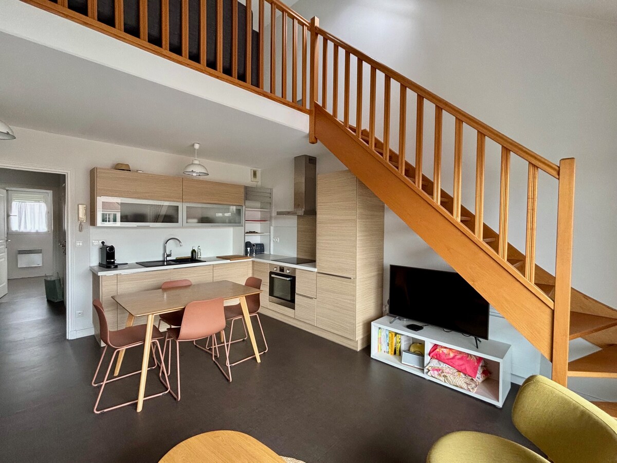 A modern kitchen and dining area are presented with light wooden cabinetry and an island counter. A dining table with four pink chairs is visible. Natural light fills the space through large windows, highlighting the warm tones of the furnishings and the staircase leading to the mezzanine.