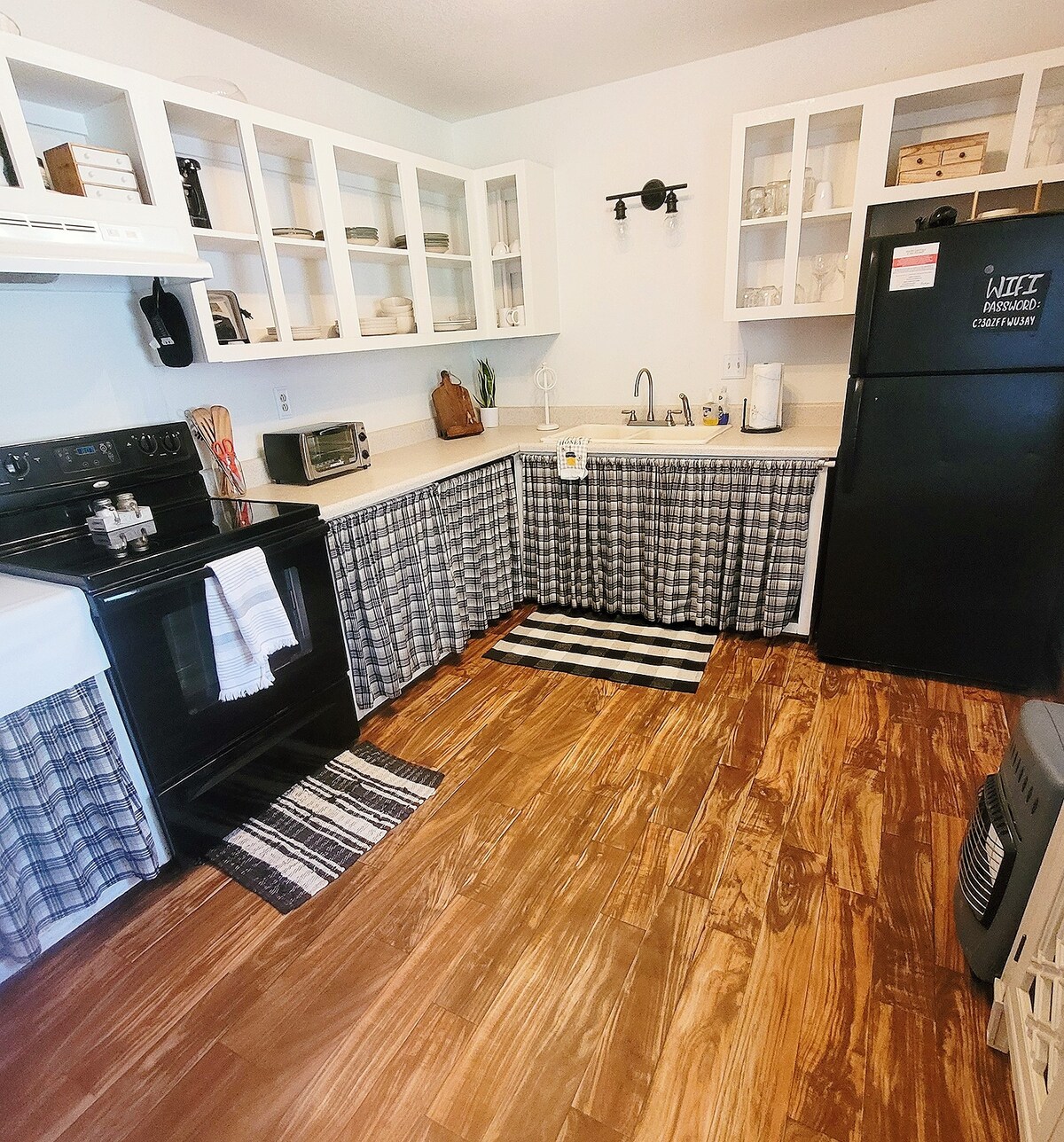 The kitchen features dark appliances, including a stove and refrigerator, paired with light cabinetry and open shelving. Checkered curtains hang under the countertop, and striped rugs add a texture contrast to the hardwood floor. Ample natural light brightens the space.