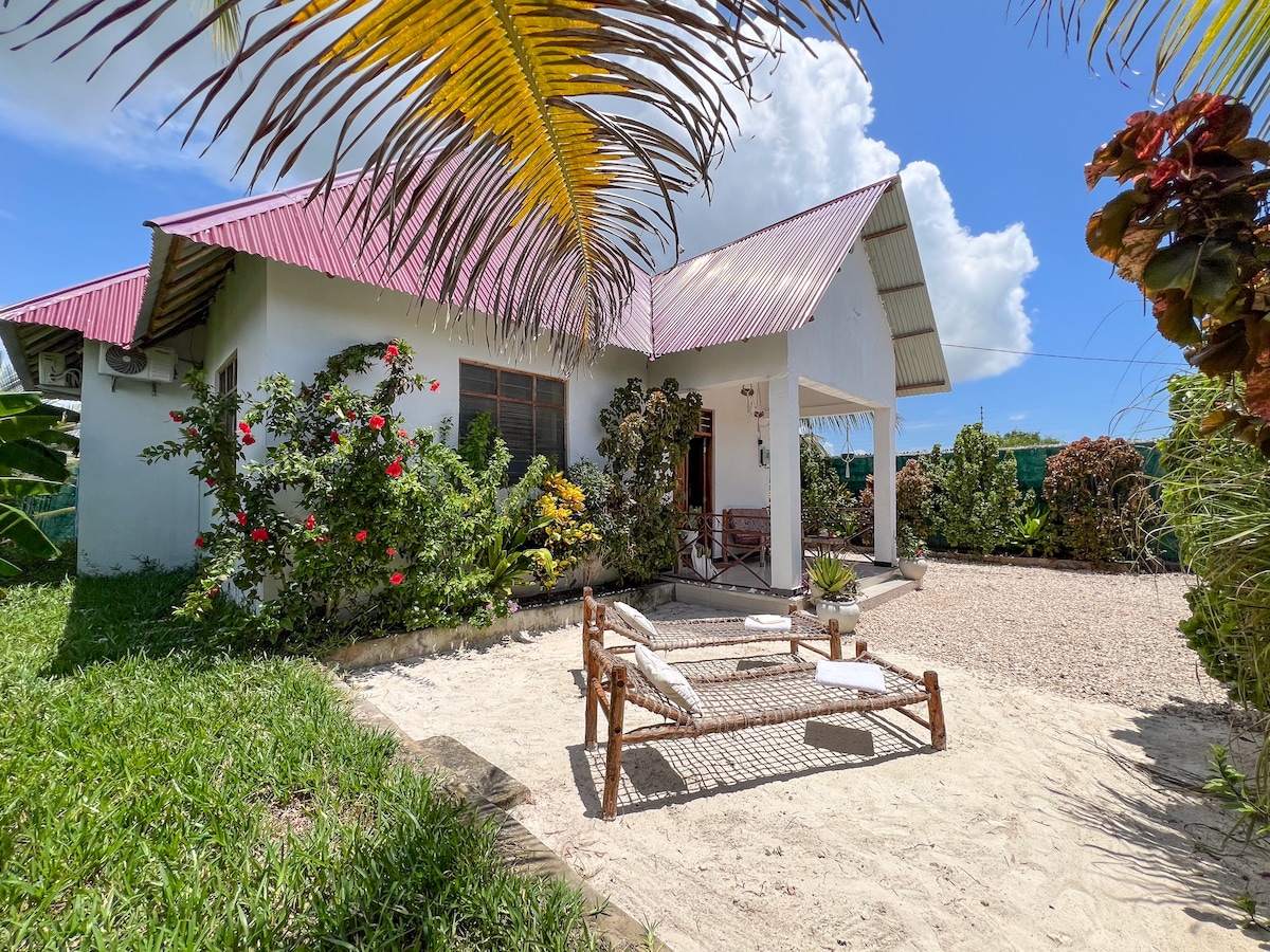 A modern house is set within a lush garden, featuring a vibrant mixture of green grass and colorful flowers. A seating area with two loungers is positioned on the sandy ground, complemented by a covered porch structure. Bright blue skies and white clouds create a serene backdrop.