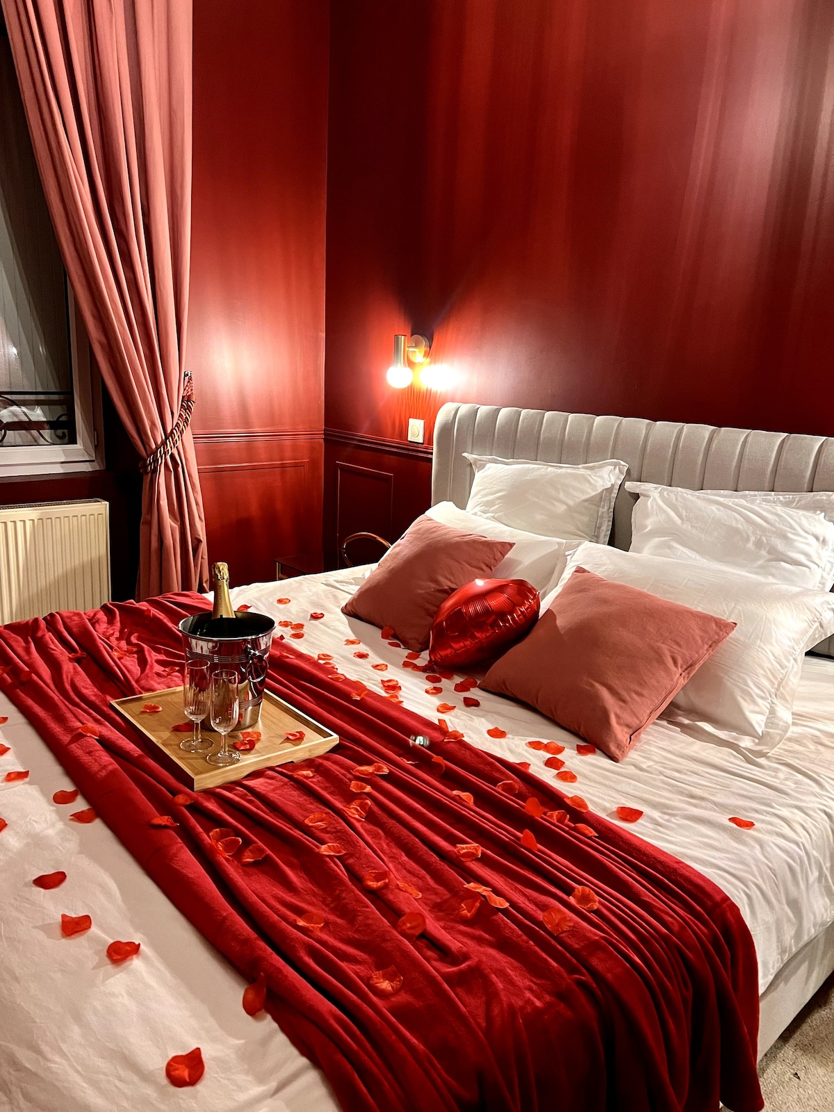 A romantic bedroom features a king-size bed adorned with white linens and plush pillows. Red rose petals are scattered across the bed, and a tray with champagne flutes and a bottle sits at the foot. Soft lighting enhances the warm red walls and curtains.