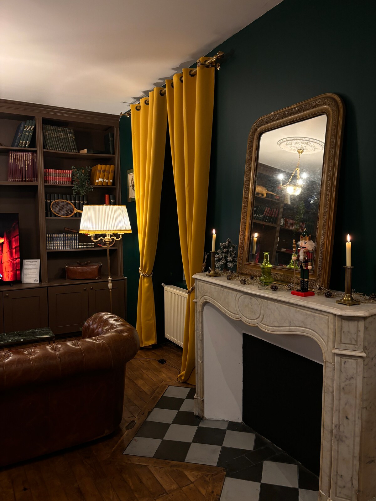 A cozy corner of the living room features a vintage-style marble fireplace with a black and white tiled hearth. A large mirror reflects soft lighting, and a yellow curtain frames a window. A welcoming leather armchair complements the bookshelf filled with books and decorative items.