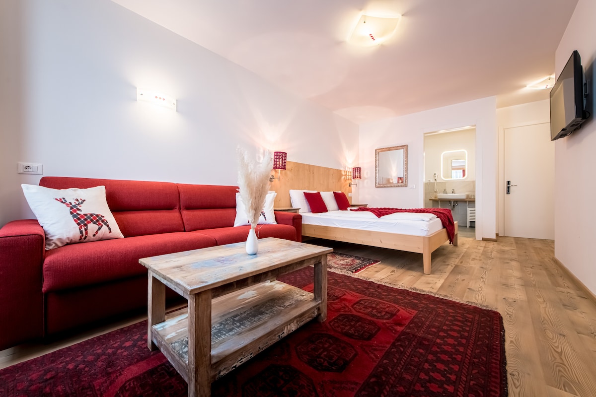 The spacious room features a red sofa and a wooden bed with a white duvet. A wooden coffee table sits on a decorative rug. A mirror is visible in the background, alongside the entrance to a private bathroom. Wall-mounted lights provide soft illumination.