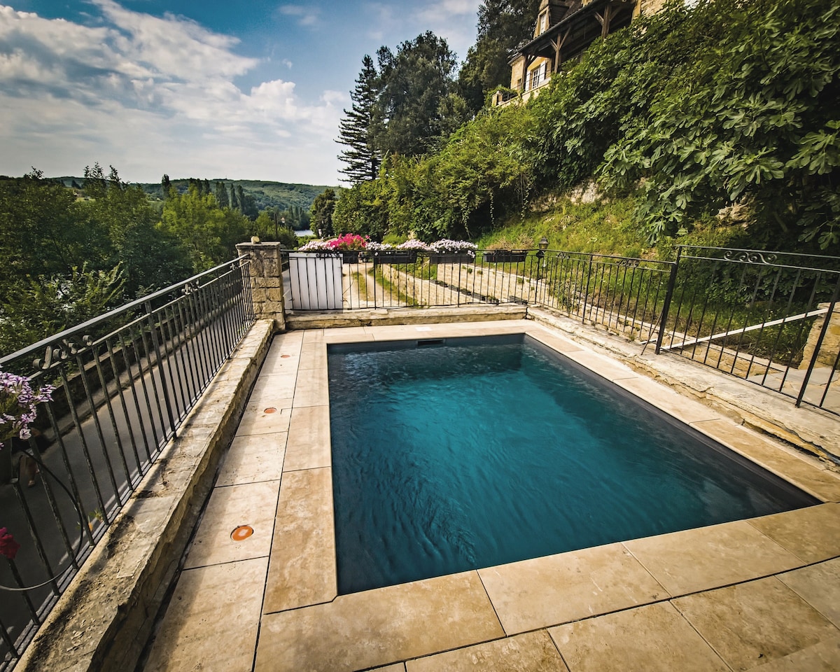 A private rectangular pool is surrounded by a stone patio, offering a clear view of the lush greenery and hills beyond. The pool area is bordered by wrought iron fencing, with colorful flower beds visible in the background, enhancing the tranquil setting.