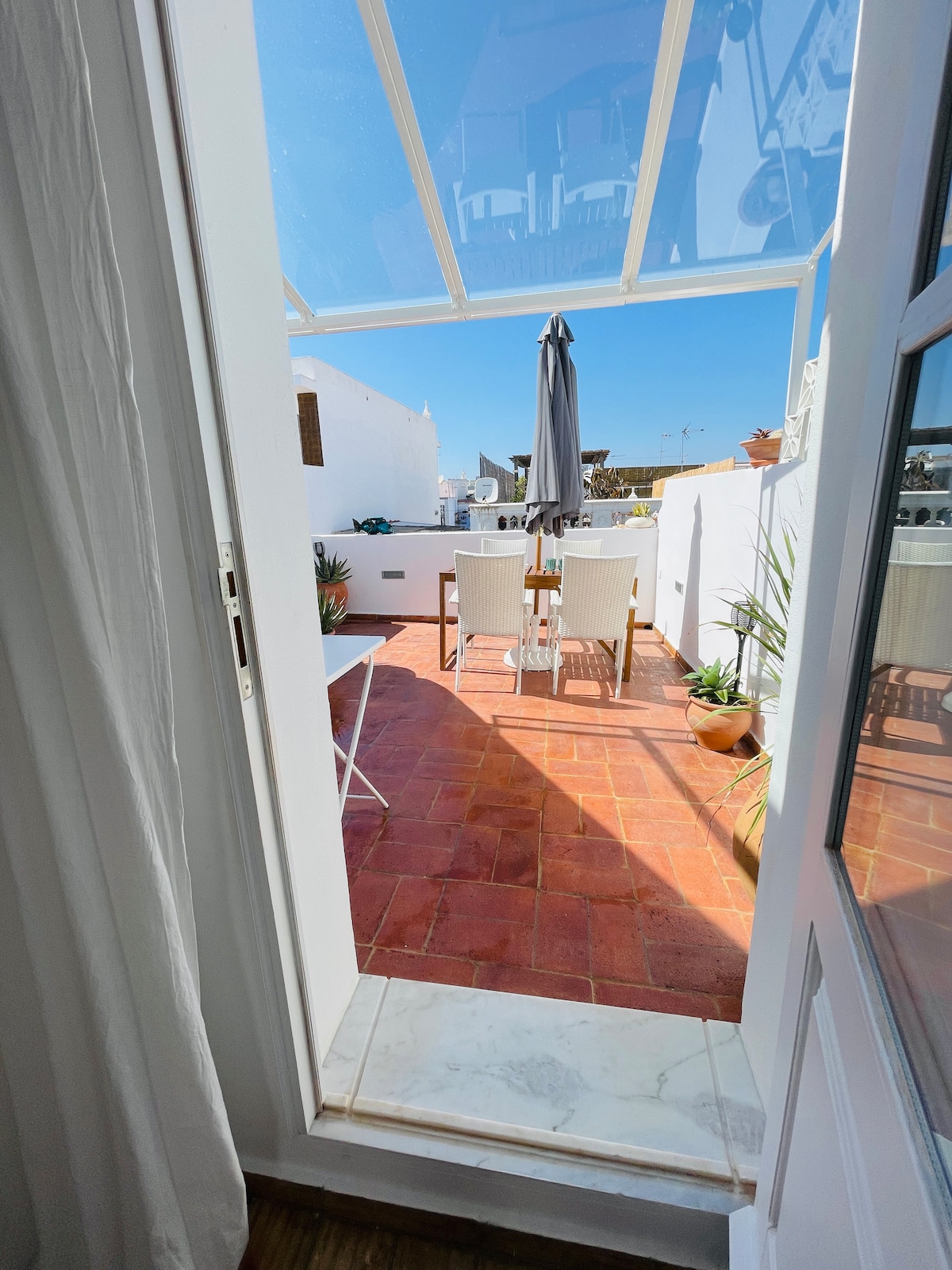 Access to a private terrace is shown, featuring a dining area with a table and four chairs, set against a backdrop of white walls and vibrant greenery. A large umbrella provides shade, while sunlight illuminates the terracotta flooring.
