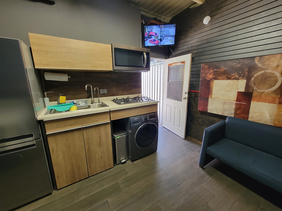A compact kitchen area features wooden cabinets, a stainless steel refrigerator, and a stove. A washing machine is positioned next to the kitchen sink. A small blue couch sits against the wall, while a flat-screen television is mounted above the counter, visible from the kitchen space.