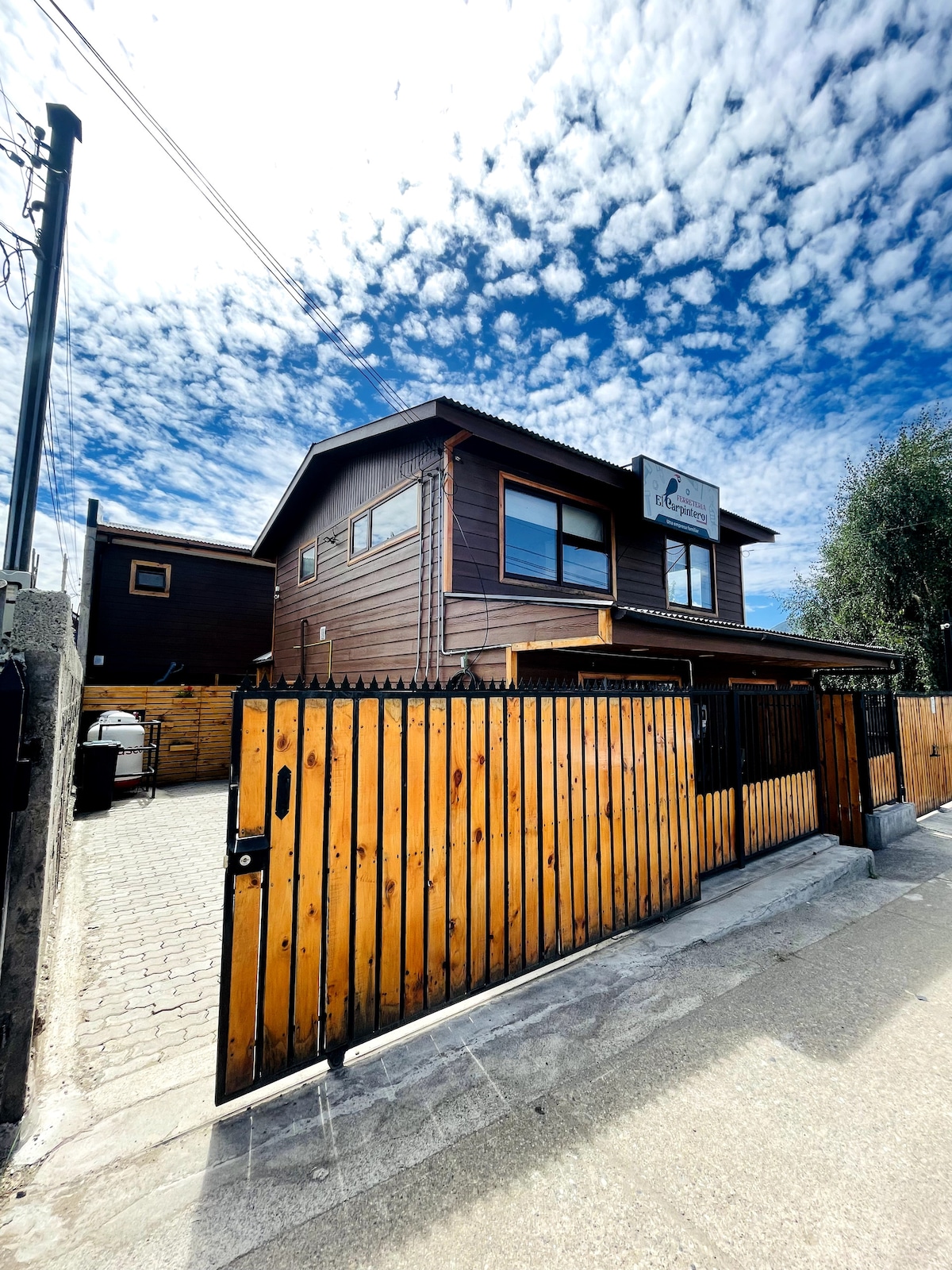 The exterior of the building showcases a two-story structure made of wood, featuring large windows that allow natural light to enter. A gated entrance provides access to a secure parking area, while the sky above is filled with white, fluffy clouds.