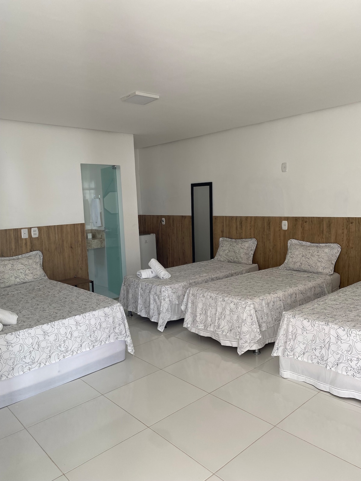 A comfortable room is arranged with three beds, each topped with light bedding featuring floral patterns. A large mirror is positioned against one wall, complementing the neutral décor of the space. Natural light fills the room through the window, contributing to a bright atmosphere.