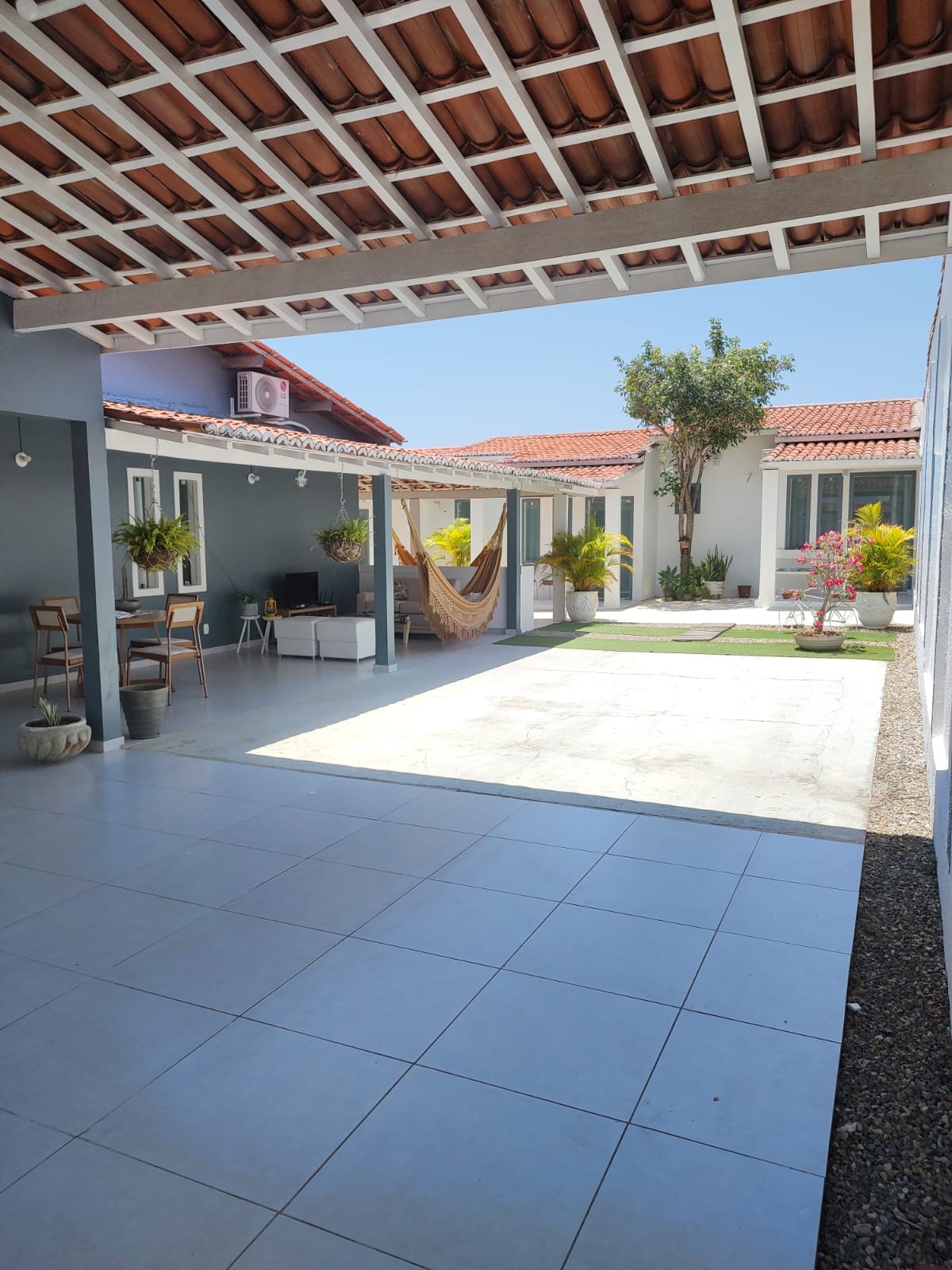 A bright outdoor area features gray tiled flooring and a shared space with seating arrangements. Potted plants add greenery around the space, while a hammock is suspended nearby. The surrounding walls are painted in light colors, creating a cheerful atmosphere under a sunny sky.