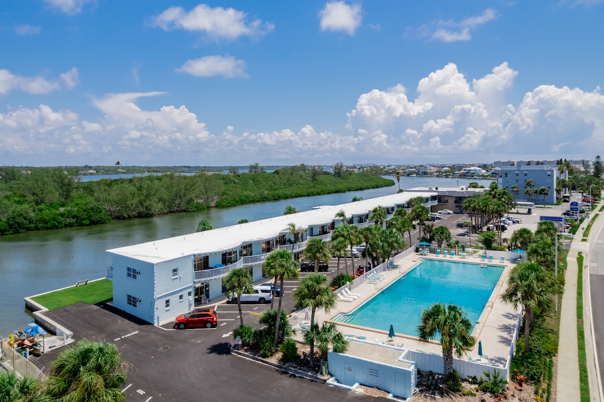 The image captures a spacious outdoor area featuring a junior Olympic-sized swimming pool surrounded by palm trees. The Intracoastal Waterway runs nearby, with a row of buildings visible along the edge. The scene showcases a clear blue sky with fluffy clouds.
