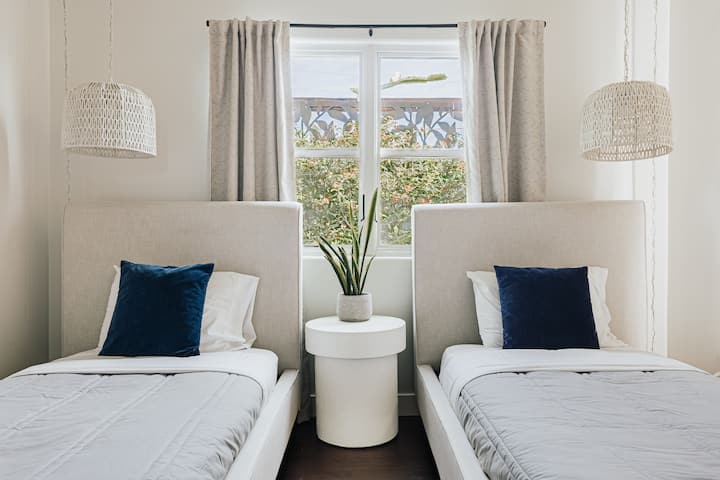 This bright bedroom features two twin beds with upholstered headboards, soft gray bedding, and navy accent pillows. Natural light flows through the window, with woven pendant lights and a modern nightstand adding a cozy, stylish touch.