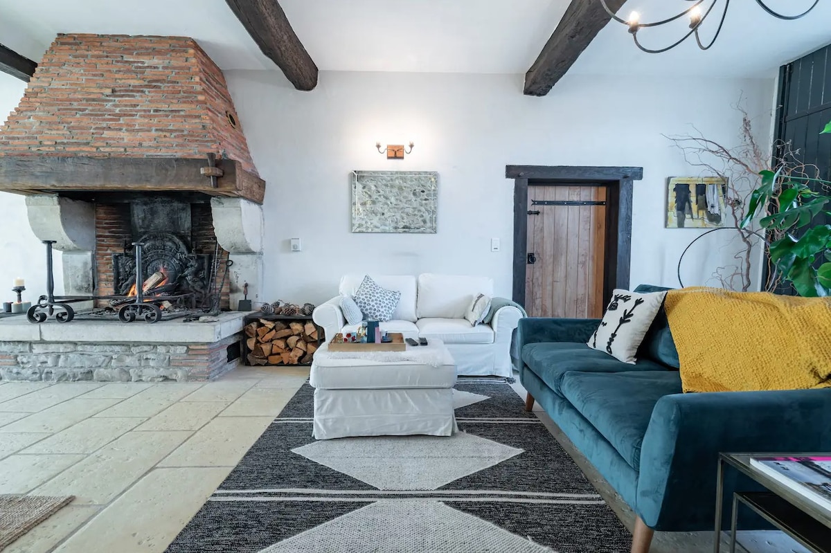 A spacious living room features exposed wooden beams and a stone fireplace, with logs stacked nearby. Seating includes a white couch and a teal sofa, complemented by a textured area rug. Artwork is displayed on the walls, providing a cozy and inviting ambiance.