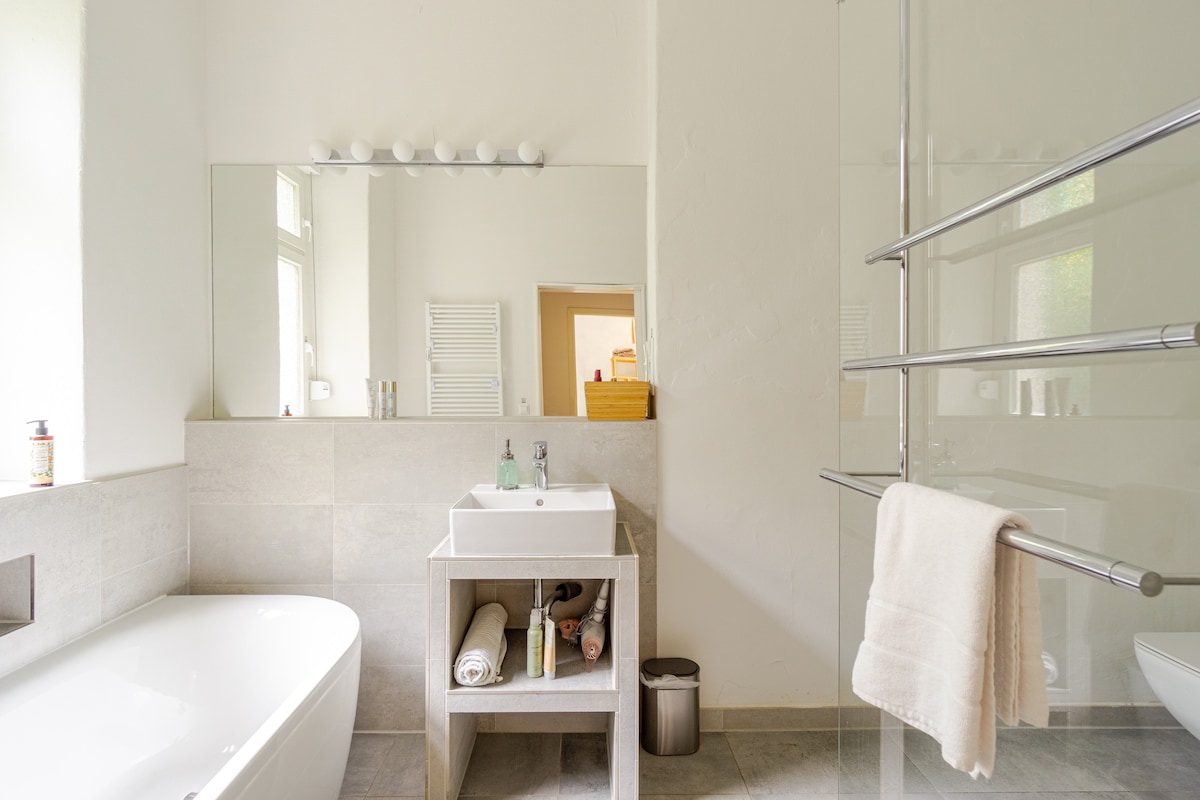 The bathroom features a large walk-in shower and a separate bathtub. A modern vanity with a sink is positioned alongside neatly organized toiletries and a towel rack. Natural light illuminates the space from the windows, creating a clean and tranquil environment.