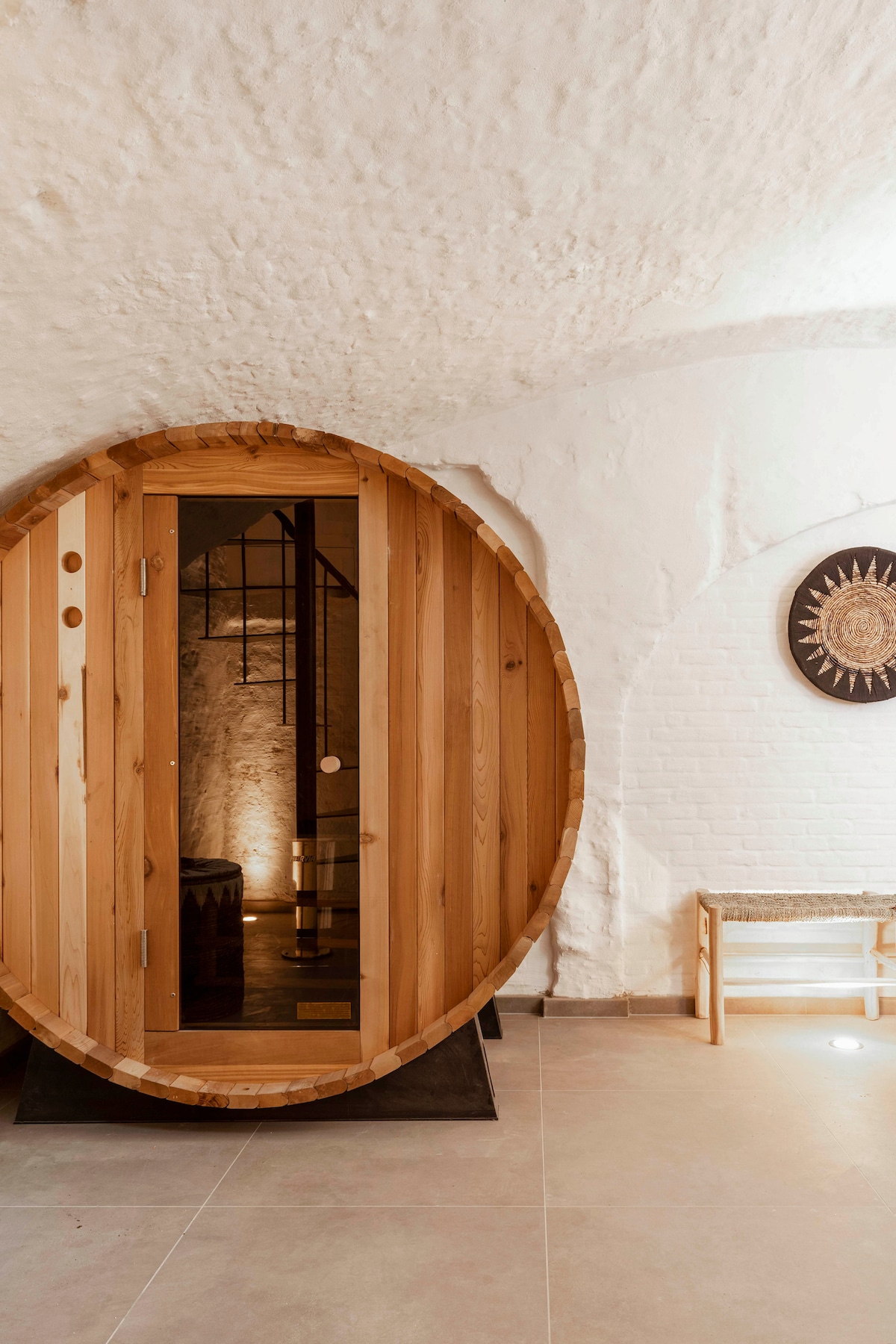 A wooden sauna hut is prominently featured, its circular shape set against a smooth white wall. Soft lighting enhances the natural wood grain, while adjacent seating and decorative wall art provide a warm and inviting backdrop. The atmosphere is calm and soothing.