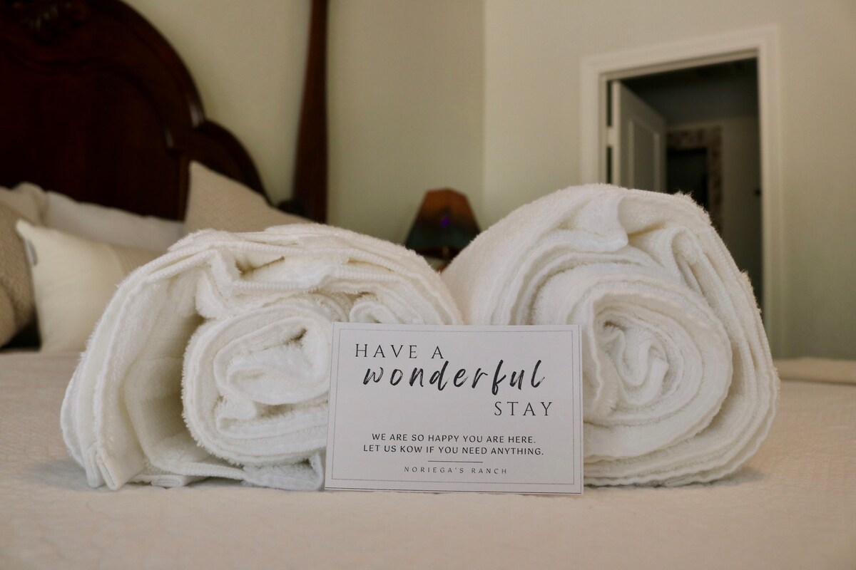 Freshly laundered white towels are neatly rolled on a bed, accompanied by a card welcoming guests. The inviting bedding and a subtle background hint at a cozy, well-maintained space. Natural light filters through the room, enhancing its calming atmosphere.