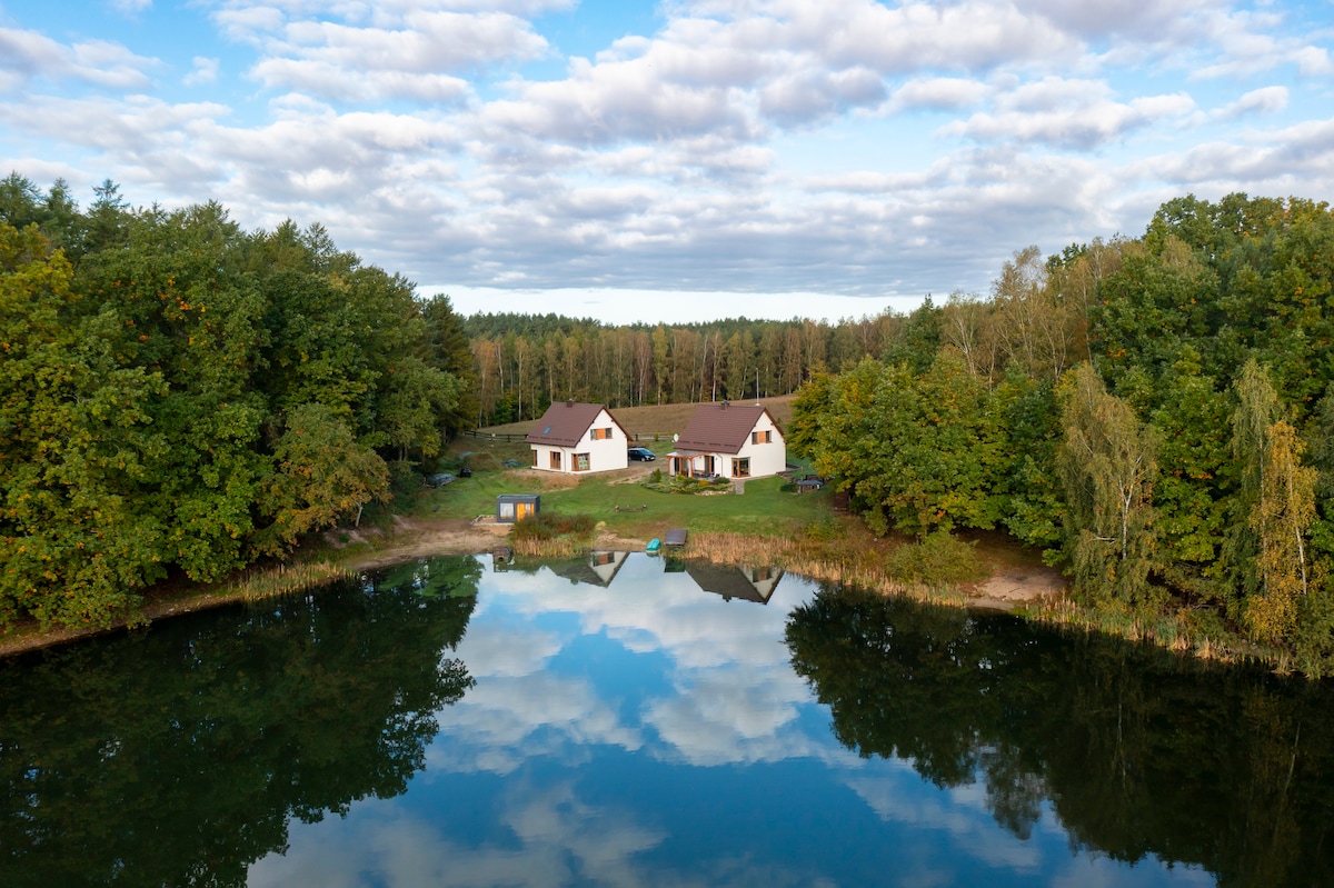 A scenic view of two charming houses is reflected in a tranquil lake. Surrounded by lush greenery and trees, the peaceful setting invites relaxation. The clear sky, dotted with fluffy clouds, enhances the serene atmosphere of this natural retreat.