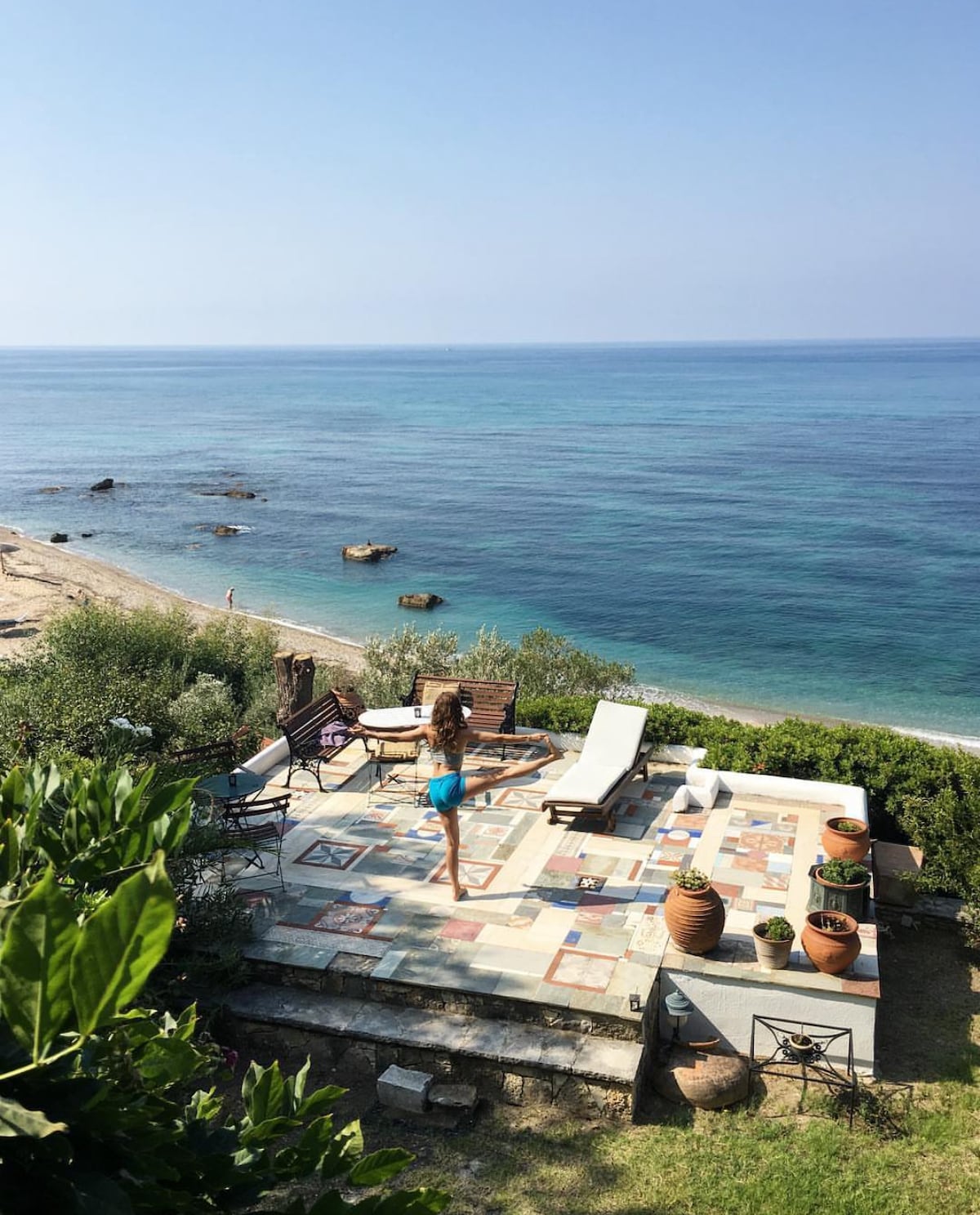 A sunlit outdoor terrace overlooks the calm blue waters of the Ionian Sea. Rustic stone tiles cover the ground, surrounded by potted plants and sturdy wooden furniture. A lounge chair is positioned for sunbathing, while olive trees provide shade in the background.