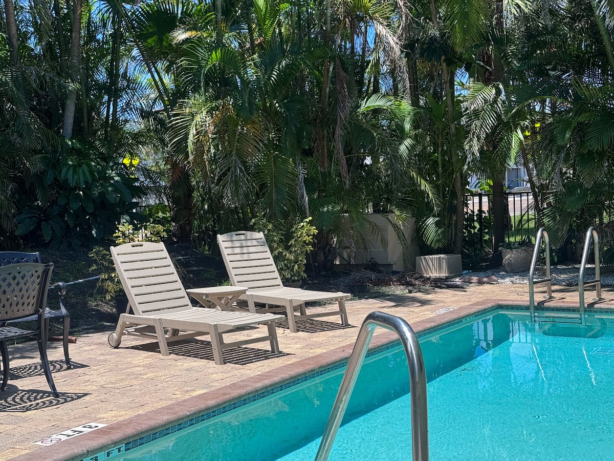 A tranquil swimming pool is surrounded by lush green landscaping. Two light-colored lounge chairs are positioned by the water's edge. A stair rail is visible at one end of the pool, providing access for guests.