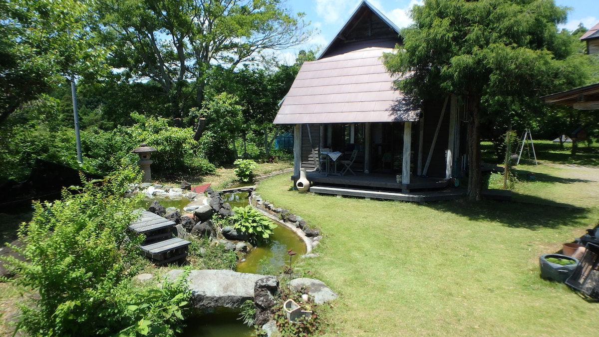 A charming cottage surrounded by lush greenery features a large garden with various plants and a serene pond. The wooden structure showcases a porch area, inviting guests to enjoy the outdoor space. Nearby, a swing set adds a playful touch to the tranquil environment.
