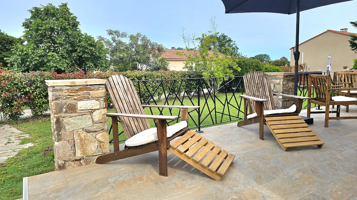 Two wooden lounge chairs with cushions are positioned on a terrace, facing a lush green garden. A large umbrella provides shade, and a stone railing adds a rustic touch to the outdoor space. Decorative flowers are seen in the background.
