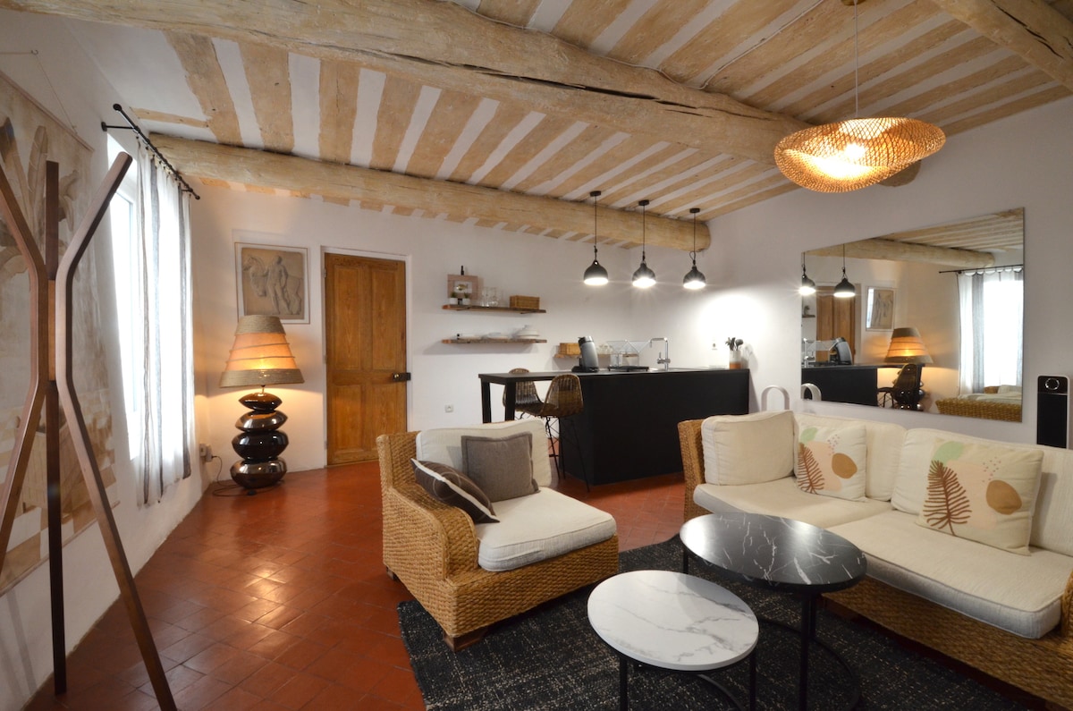 A cozy living space features a comfortable seating area with a woven sofa and a marble-top coffee table. Exposed wooden beams adorn the ceiling, and stylish lighting fixtures hang above. A dark kitchen counter is visible in the background, along with a large mirror enhancing the room's brightness.