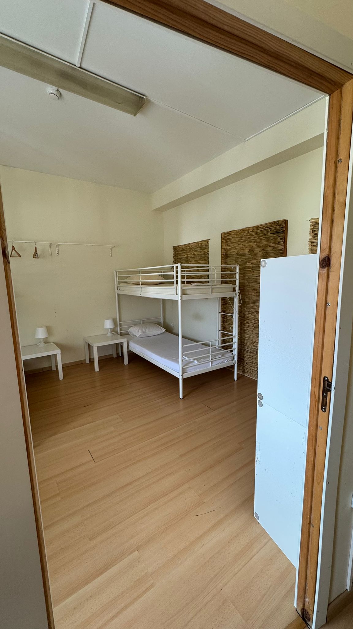 A bright hostel room is viewed through an open door. It contains a bunk bed with an unmade top bunk, two bedside tables, and a wardrobe. Natural light fills the space, highlighting the smooth wooden flooring and neutral-colored walls.