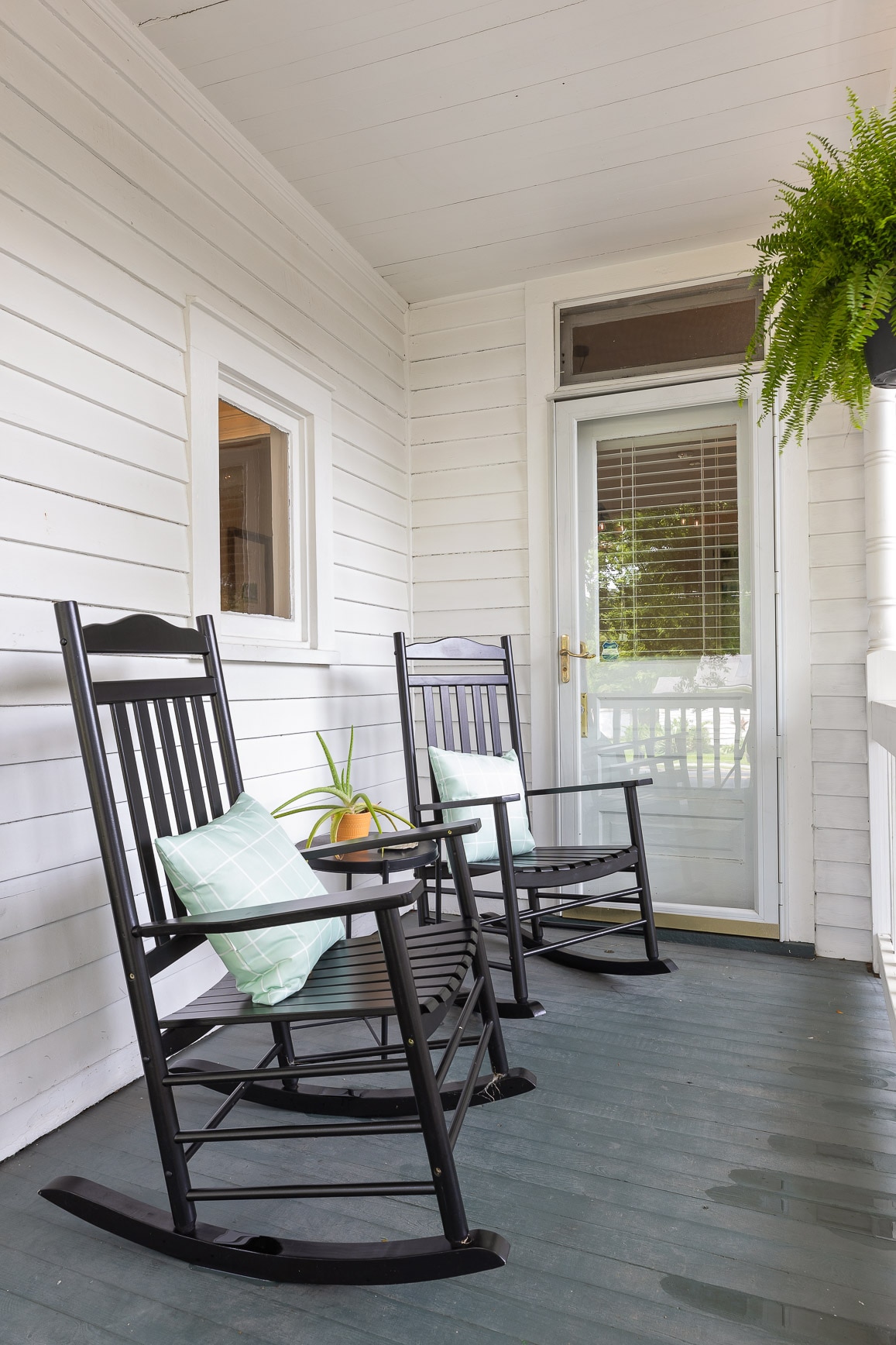 A welcoming front porch is showcased with two black rocking chairs, each adorned with a light green cushion. A potted plant adds a touch of greenery, while sunlight filters through a door leading indoors.