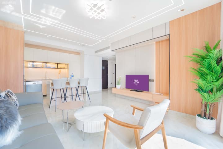 Intérieur blanc éclatant avec un canapé gris, un téléviseur élégant et un coin repas moderne. Les accents de bois clair rehaussent l'ambiance fraîche et aérée, créant un espace de vie propre et contemporain.