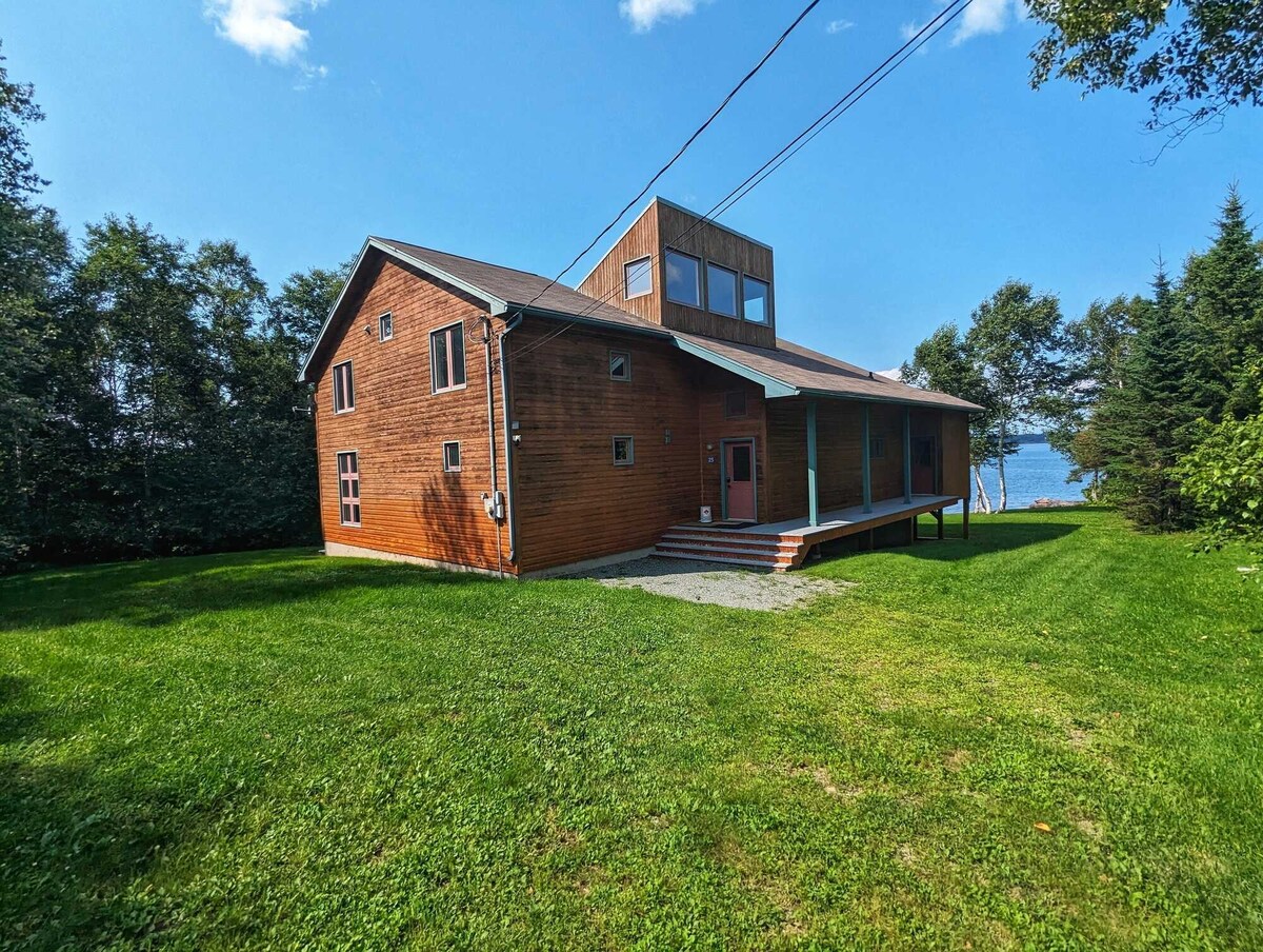 A spacious three-story wooden house is depicted, featuring large windows that provide views of the surrounding landscape. A well-kept lawn surrounds the building, with a path leading to the entrance. Trees are visible in the background, adding to the natural setting.