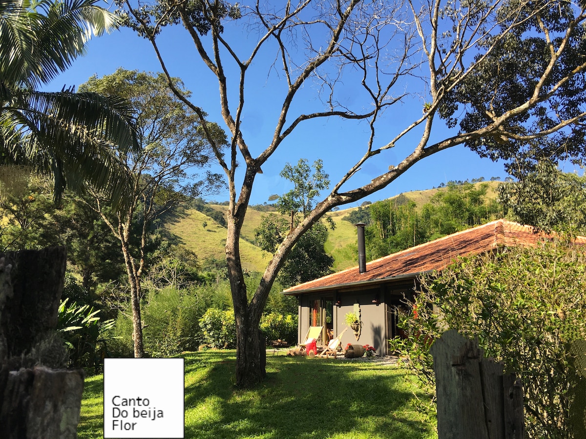 The cozy exterior of the Canto do Beija Flor is framed by lush greenery and towering trees. A rustic structure with a clay-tile roof reflects a harmonious blend with the natural surroundings. The bright sky enhances the tranquil atmosphere of the peaceful garden setting.