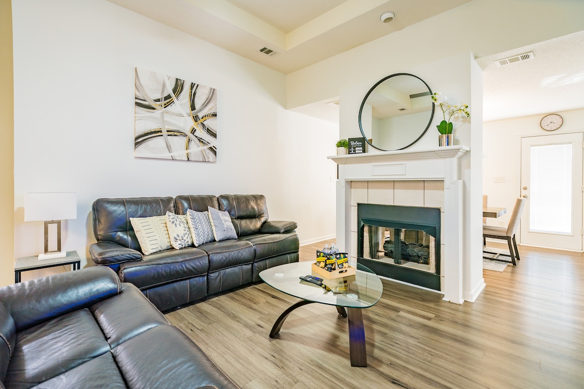 The living room features a cozy seating arrangement with dark leather sofas and a glass coffee table. A decorative mirror hangs above a fireplace, while a piece of abstract wall art adds visual interest. Natural light filters in through the door leading to the outside.
