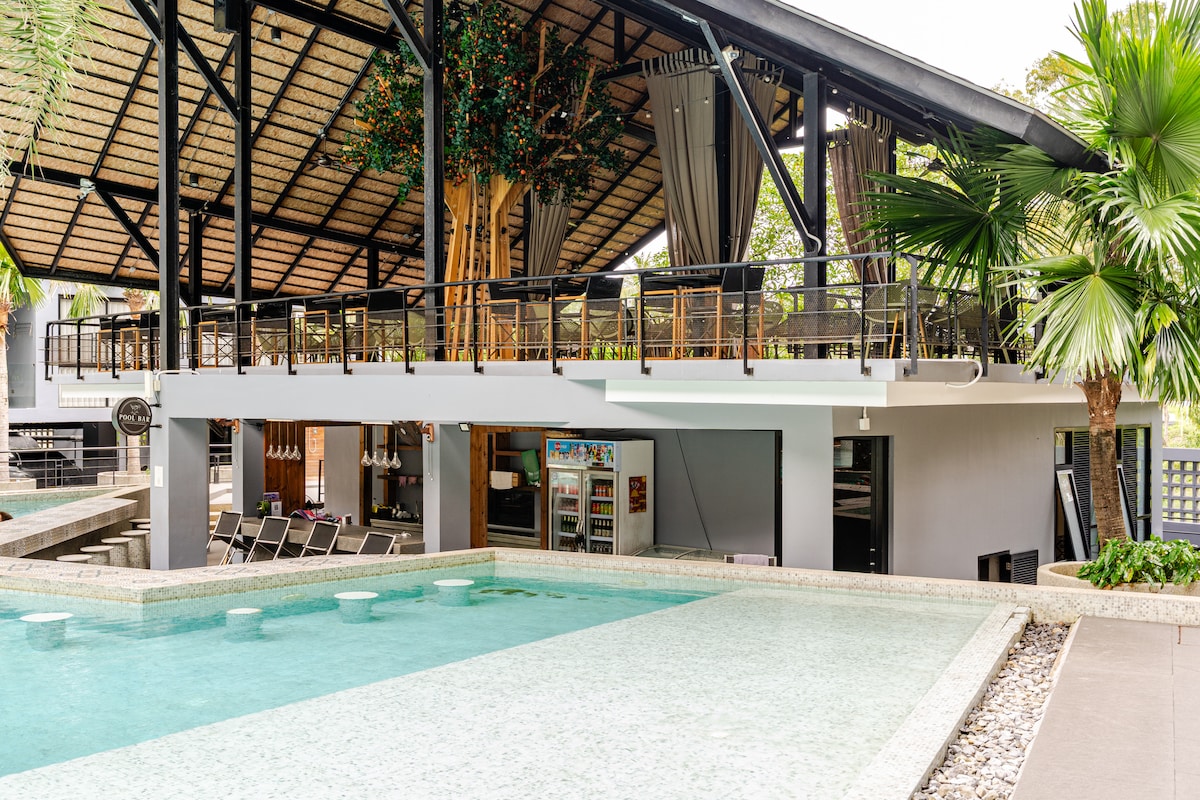 A modern rooftop infinity pool is surrounded by a sleek wooden deck and tropical palm trees. The pool's clear water reflects the nearby shaded seating area, which includes loungers and a bar area. Overhead, fabric drapes create a relaxed ambiance in this inviting outdoor space.