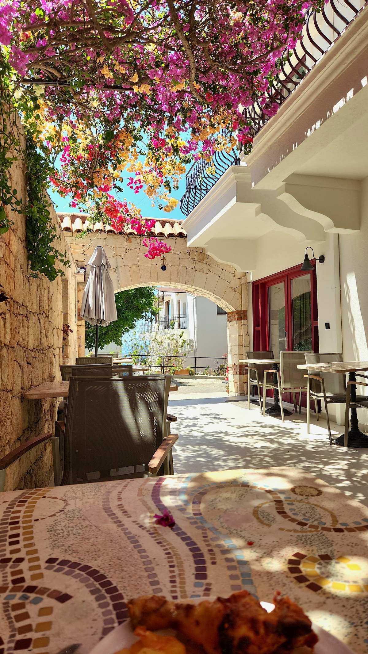 An outdoor seating area is presented, featuring a stone archway adorned with blooming bougainvillea. Light filters through the colorful flowers, creating a vibrant atmosphere. Tables and chairs are arranged on a patterned surface, enhancing the inviting space for relaxation.