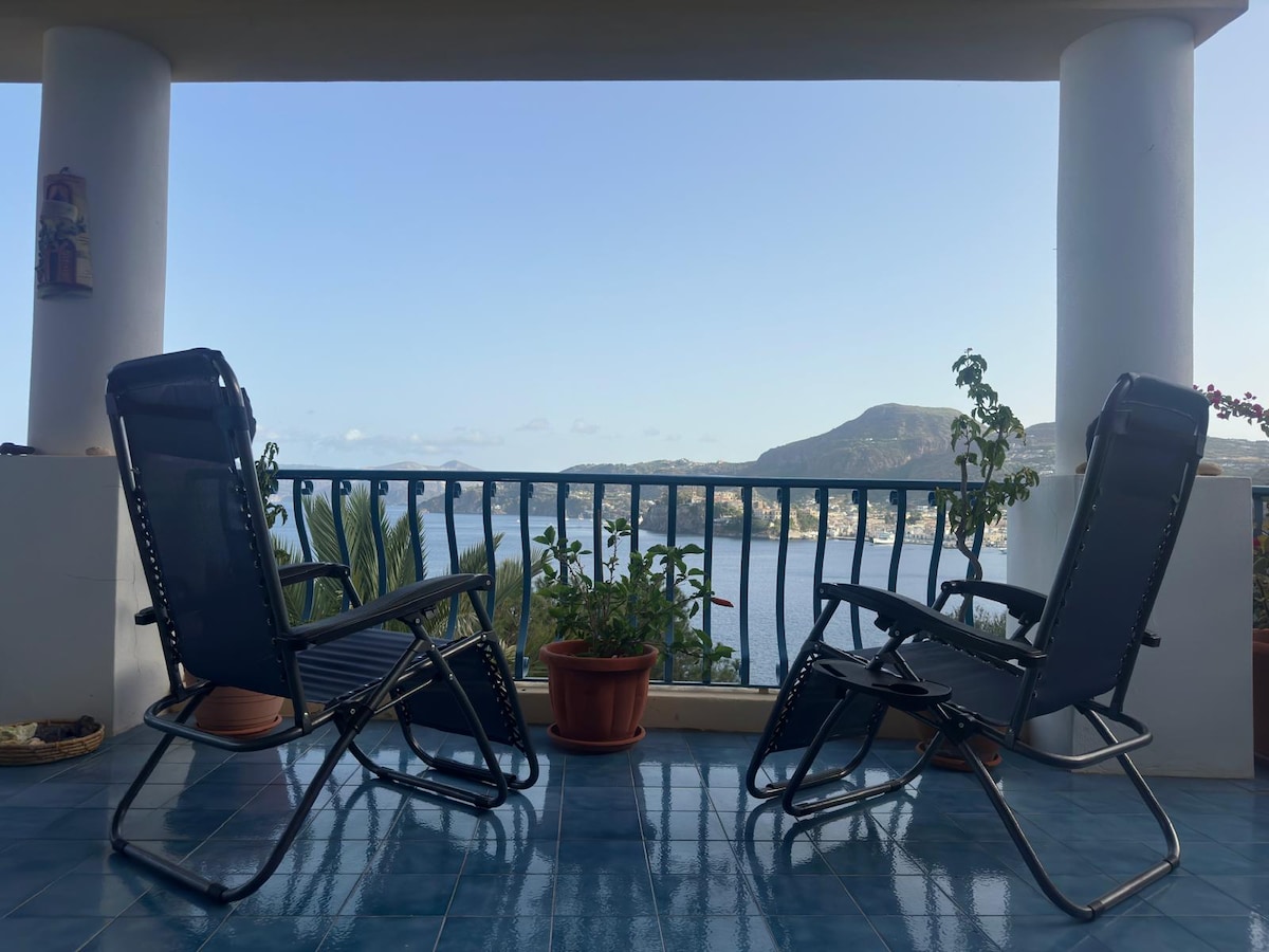Two reclining chairs are positioned on a spacious terrace, offering a clear view of the serene bay and mountainous landscape. Potted plants add a touch of greenery, while the blue tiled floor complements the coastal scenery.