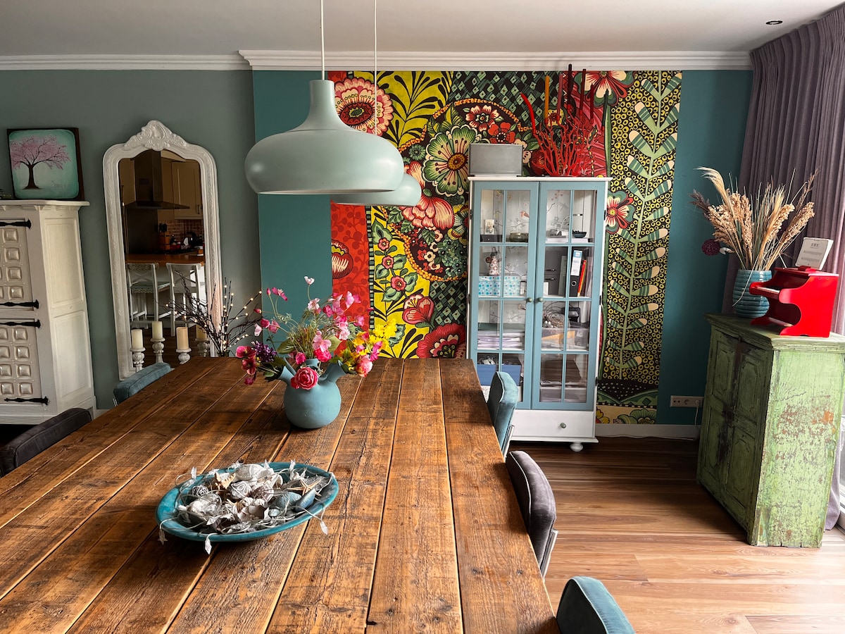 A spacious dining area features a large wooden table at its center, surrounded by comfortable chairs. A vibrant floral accent wall adds a burst of color, while a cabinet showcases decorative items. Soft natural light fills the room, enhancing the inviting atmosphere.