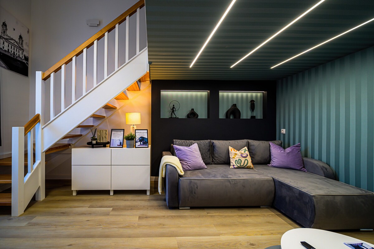 A spacious living area is showcased, featuring a sectional sofa with purple cushions. A minimalist white cabinet holds decorative items, while subtle lighting enhances the modern design. The staircase leads to the upper level, and walls display soft green stripes, contributing to the overall aesthetic.