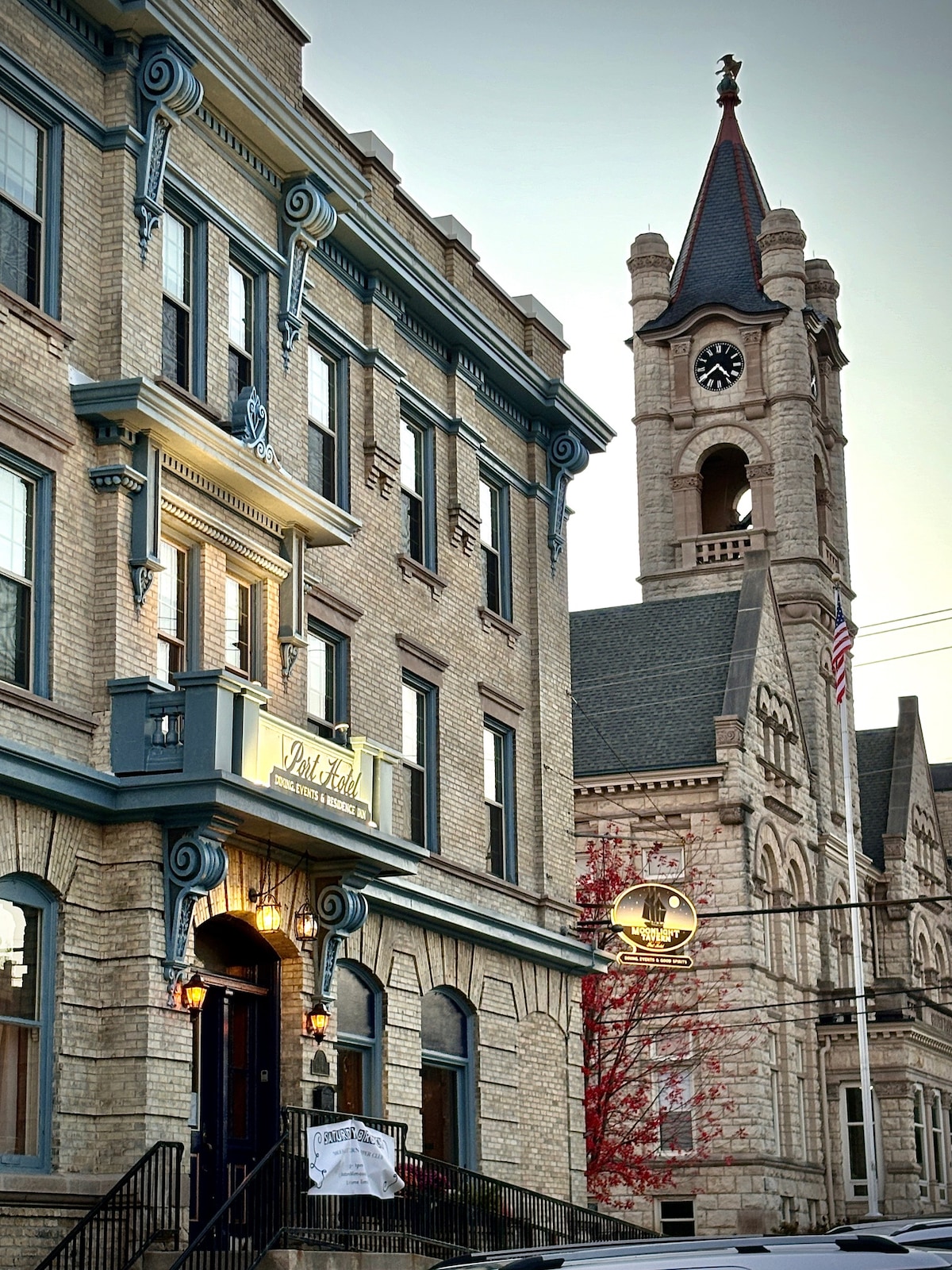 The exterior of the historic Port Hotel is captured, showcasing intricate architectural details and a welcoming entrance. A clock tower rises in the background, adding to the charm of the building's vintage style. The image also includes decorative elements and surrounding trees hinting at seasonal changes.
