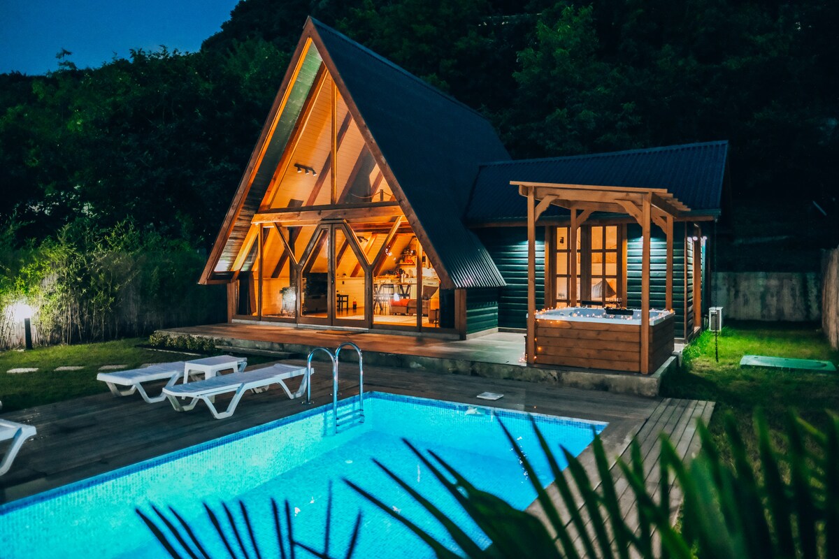 A two-story cabin features a steeply pitched roof and large windows, allowing for natural light to fill the interior. A refreshing pool is depicted in front, accompanied by lounge chairs, while a wooden gazebo houses a hot tub beside the cabin.