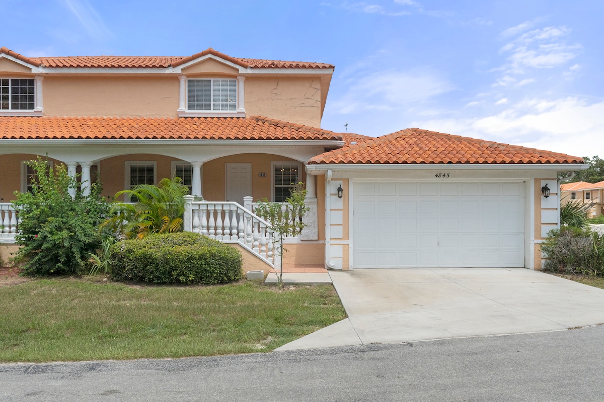 A two-story villa is showcased, featuring a warm clay tile roof and front porch with decorative railings. The well-maintained lawn includes shrubs and plants, while a two-car garage is visible on the right side of the image, providing ample parking space.