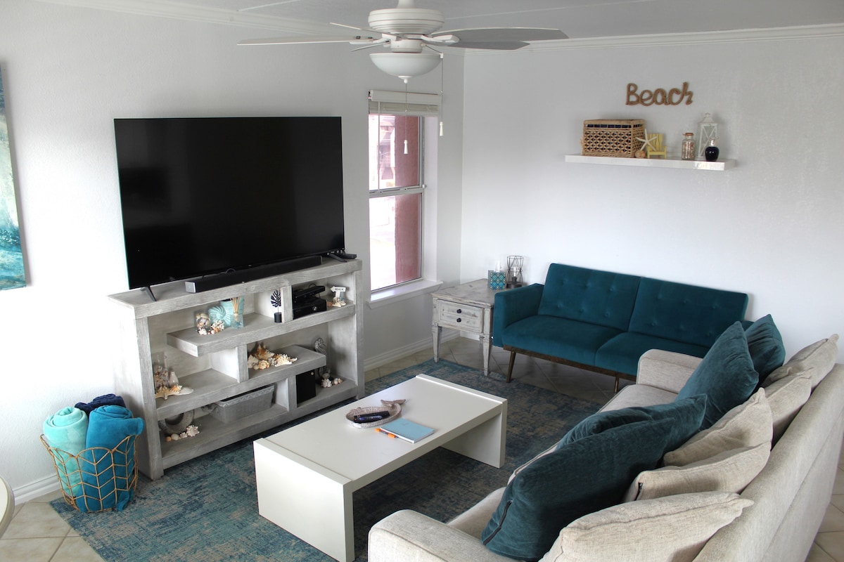 A cozy living room is featured, showcasing a large flat-screen television mounted on a light-coloured shelf. A comfortable sofa in neutral tones is paired with a teal accent chair. Decorative elements include beach-themed décor and a patterned area rug.