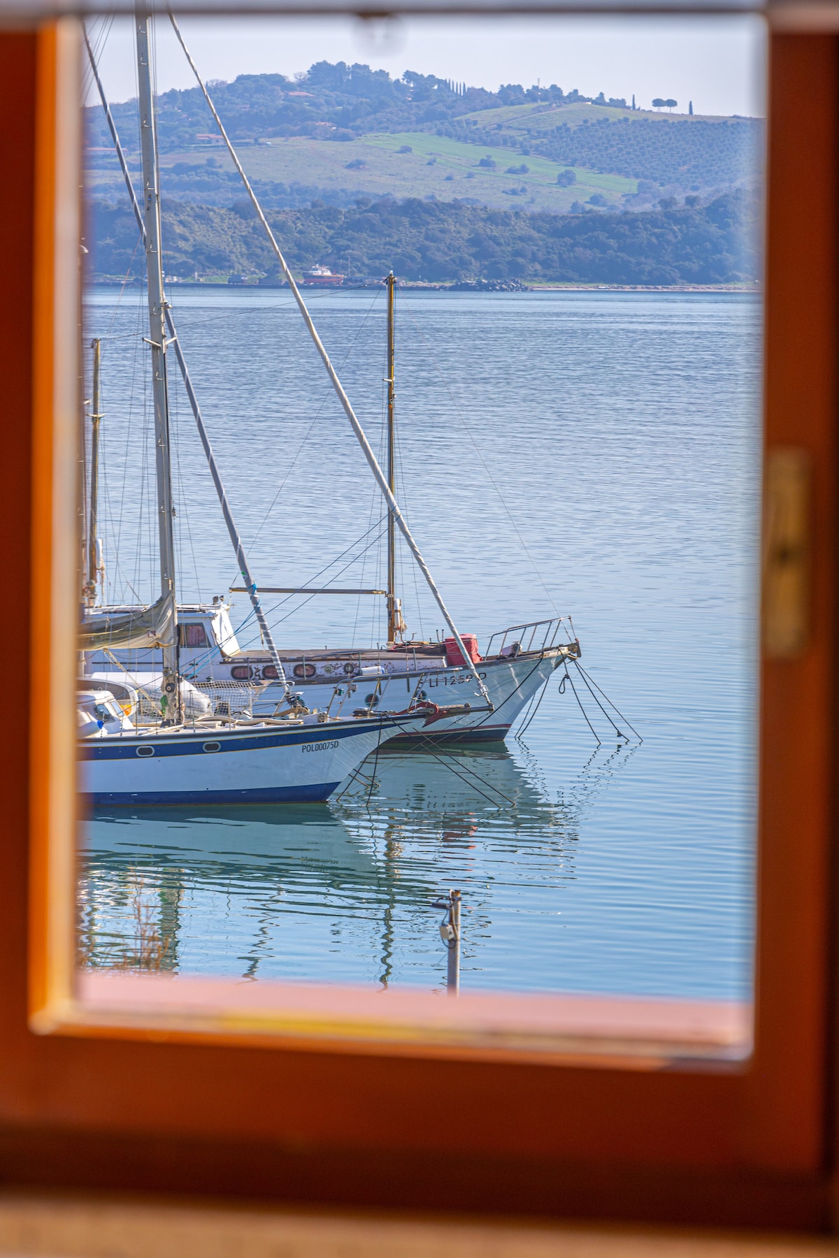 A serene view is framed by a wooden window, revealing a calm body of water with a sailboat gently moored. Lush green hills are visible in the background, reflecting the tranquility of the surroundings.