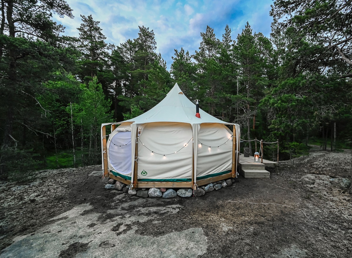 A Lotus Belle glamping tent is situated among lush greenery, surrounded by tall trees. The structured canvas features a conical top and is partially illuminated by string lights. A wooden deck leads to the entrance, enhancing accessibility and connection with the natural surroundings.