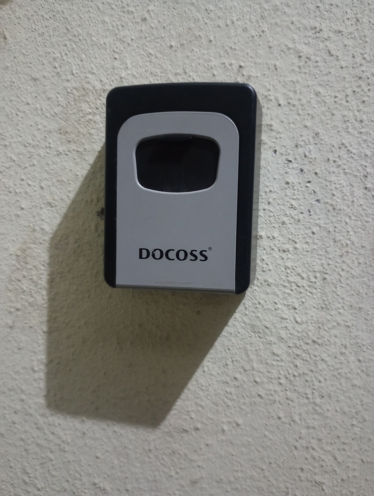 A device is mounted on a textured wall, featuring a dark casing with a clear viewing window displaying the brand name 'DOCOSS'. A shadow is cast on the wall from the device, highlighting its rectangular shape.