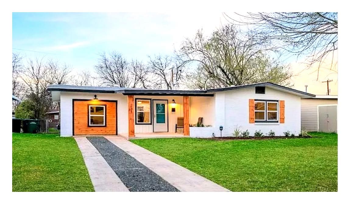 A single-story home is presented with a well-maintained lawn and a gravel pathway leading to the entrance. Exposed wooden accents frame the front windows and door, while the garage features a charming wooden door. A clear sky and tree silhouettes create a serene backdrop.
