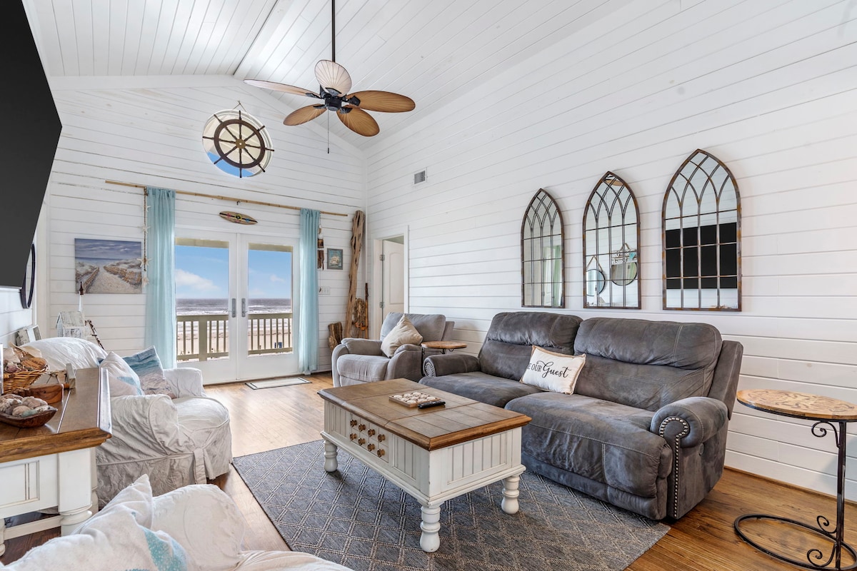 A bright living room features large windows with a view of the beach, allowing natural light to fill the space. Comfortable seating is arranged around a central coffee table, and decorative elements include a ceiling fan and unique wall mirrors that enhance the coastal aesthetic.